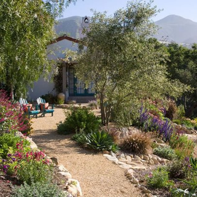Scenic view of home with mountains in background, porch and decomposed granite walkway surrounded by trees, flowers and rock border.